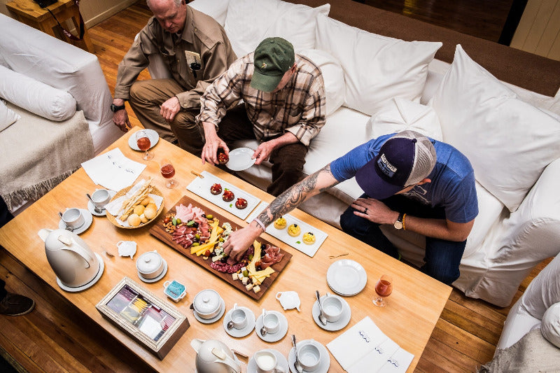 Three anglers around a wooden coffee table with food and drinks in a living room setting.
